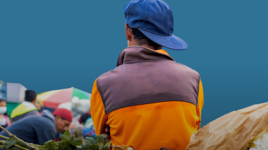 ragazzo ripreso di spalle con giacca gialla e cappellino blu e sull sfondo sacchi con alimenti